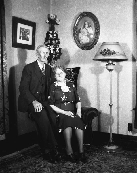 Indoor portrait of Alexander and Florentina Krueger. Florentina is sitting in a sofa chair while Alexander is sitting next to her on the arm of the chair. A lamp is next to them, and several portriats and figurines are hanging on the walls behind them.