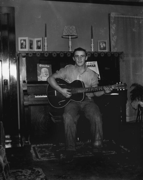 Emil Hertz sitting on a piano bench strumming a guitar in the Krueger home. Several portraits, a lamp, and two candles are on top of the piano.