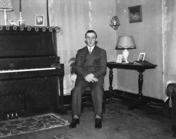 Emil Hertz in a suit sitting next to the piano in the Krueger's living room. Several portraits are on top of the piano and an end table.
