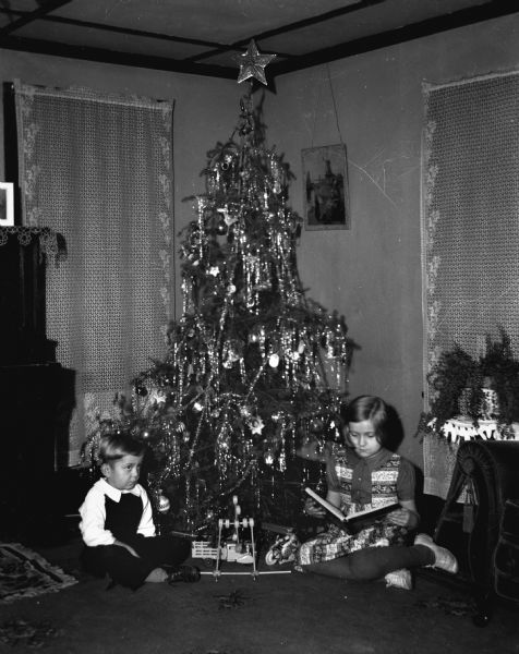 Robert and Shirley Krueger sitting in front of a decorated Christmas tree. Robert is sitting crossed legged while Shirley is sitting and reading a book. Several of their toys are sitting in between them.