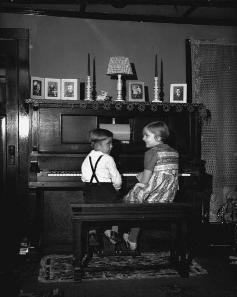 Robert and Shirley Krueger sitting on the piano bench looking at each other while playing piano together in the living room. Several portraits and other decorations are sitting on top of the piano. A decorated Christmas tree is on the right.
