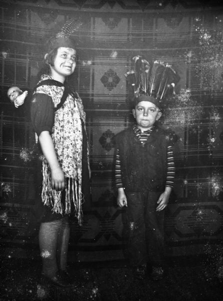 Shirley and Robert Krueger dressed up in feather headdresses as Native American Indians, standing in front of a patterned blanket backdrop. Shirley is wearing face makeup and a shawl while carrying a baby doll strapped to her back.