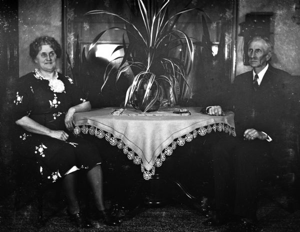 Florentina and Alexander Krueger sitting at either end of the dining room table. A tablecloth and a large potted plant are on the table.