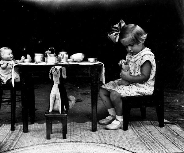 Shirley Krueger sitting outside at a small table with chairs having a tea party with several of her toys. Shirley is sitting in a chair playing with the small stuffed bunny in her lap, while her baby doll and a stuffed dog are sitting in two other chairs. A tea set with a table cloth is sitting on top of the table. A carpet has been laid out on the ground.