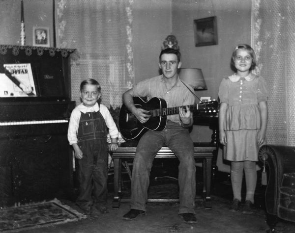 Emil Hertz playing guitar while sitting on a piano bench set in the middle of the Krueger living room. Shirley and Robert Krueger are standing on either side of him.