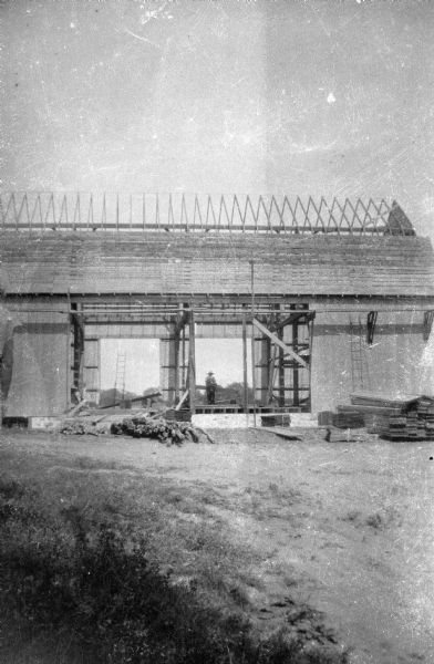The framework of a new barn being constructed. The roof of the barn is partially constructed and the doors of the barn are not yet in place. A man is standing inside the barn holding a piece of timber. Several piles of timber are stacked on the ground in front of the building.
