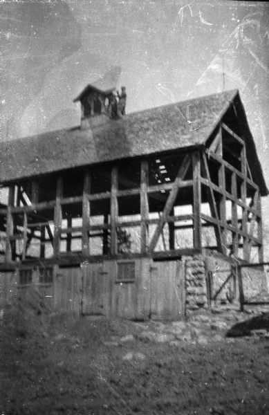 View from below of an old barn. Partially deconstructed, with only the roof, base, and framework intact. A man is standing on top of the roof removing the cupola.