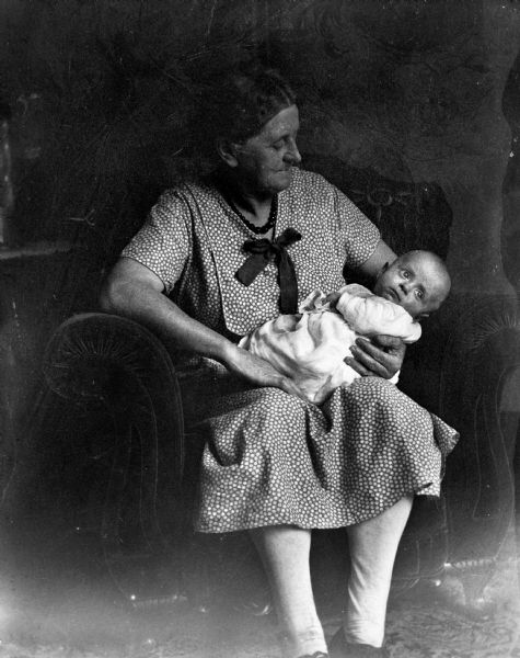 Florentina Krueger sitting in a sofa chair holding, and looking down at baby Robert Krueger, her grandson, in her lap.