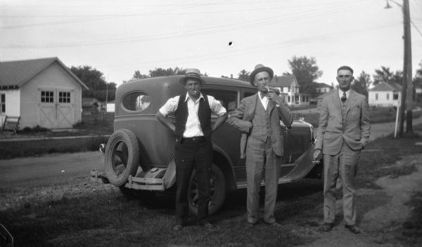 Edgar Krueger standing next to a vehicle parked along the road with two friends, smoking cigarrettes. Several houses and garages are in the background.
