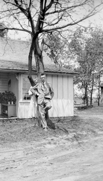 View across dirt road towards a man leaning against a tree next to the home of Edgar Krueger. He has a cigarette or cigar in his mouth. Elna Krueger is standing behind the tree peering between its two trunks over the mans' shoulder.