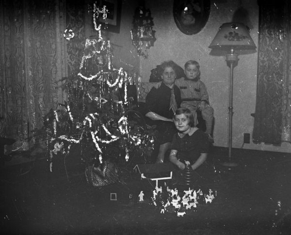 Three people sitting indoors next to a Christmas tree. Florentina Krueger is sitting in a sofa chair, and her grandson, Robert Krueger, sits on her lap. Florentina's granddaughter, Shirley, sits on the floor in front of her. A toy barn with a silo and various farm animal figures sits on the floor in front of Shirley.