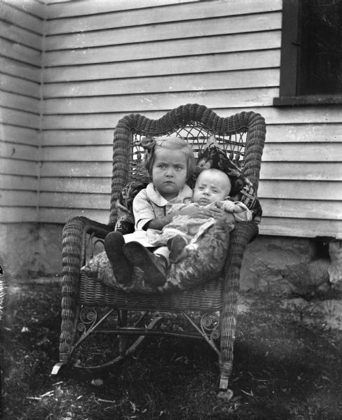 Outdoor portrait of Shirley Krueger sitting in a wicker rocking chair holding her infant brother, Robert Krueger, in her lap. The side of a house, with a window on the right, is behind them.