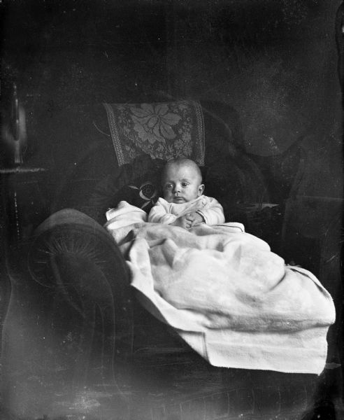 Indoor portrait of infant Robert Krueger propped up in a sofa chair with pillows. A blanket is covering him from the waist down.