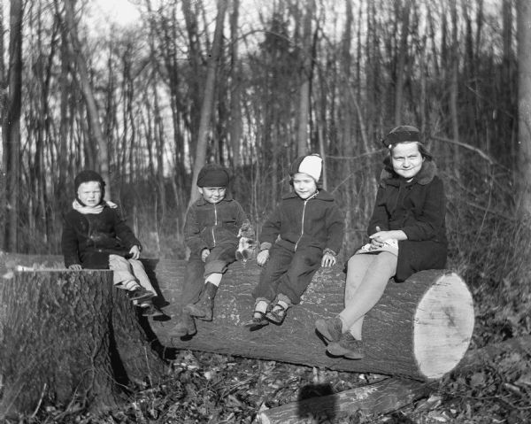 Robert and Shirley Krueger sitting on a large tree log with a young girl sitting between them. On the left another child is sitting on a large tree stump. Robert has his arm around a squirrel that is propped up next to him.