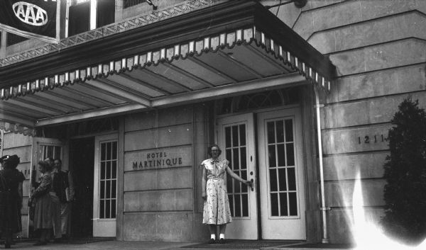 View across sidewalk towards a young woman opening the entrance door to the Hotel Martinique. Several other people are exiting from the other set of doors on the left.