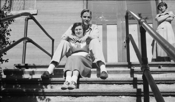 A young man sits on the top step of an entrance to a building with his arms wrapped around the shoulders of a young woman who sits on the step below him between his legs. Another young woman stands to the right of the entrance door.