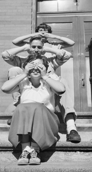 Group portrait of three young people sitting in a row on steps leading up the entrance of a building. The woman in the foreground sits on the second highest step covering her eyes with her hands. Behind her, a young man sits on the top step covering his ears, while a young woman stands behind him covering her mouth with her hands.