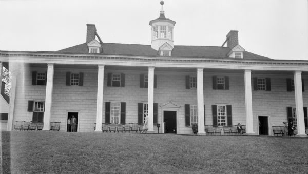 View across lawn towards the back side of George Washington's Mount Vernon. Several people are sitting and standing on the piazza. There is a cupola on the roof.