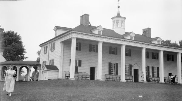 View across lawn towards the back of George Washington's Mount Vernon from the left. A woman is walking on the lawn in the left foreground, and behind her is a colonnade. Groups of other people are on the piazza. There is a cupola on the roof with a weather vane.