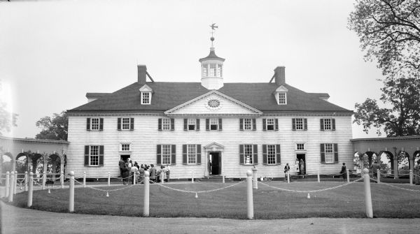 Front view of George Washington's Mount Vernon from the circular driveway. A line of people are waiting at the entrance. The cupola on the roof has a weather vane depicting a dove with an olive branch. Colonnades on the left and right connect to the servants' hall and kitchen. The Potomac River is in the background.