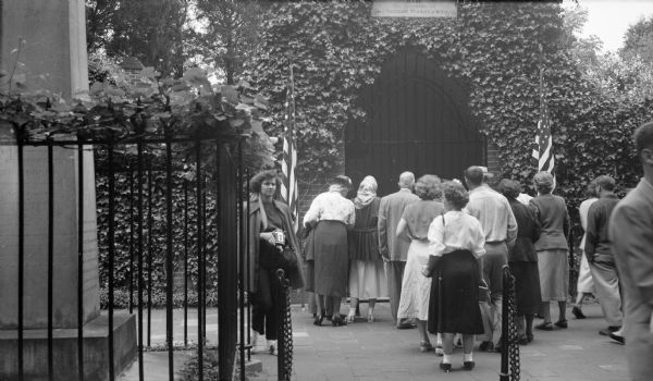 Group of people standing in front of George and Martha Washington's tombs. One woman on the left is walking away from the tomb holding a camera. Two American flags flank the sides of the gated entrance of the tomb. On the left behind the iron fence is a marble sarcophagi.