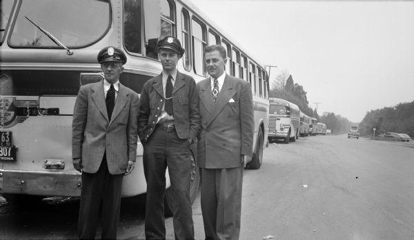 A young man posing with two bus drivers in front of a row of buses parked on the side of the road. Several vehicles are parked in a parking lot across the road on the right.