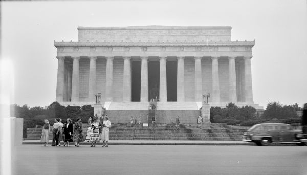 View of the Lincoln Memorial from across Lincoln Memorial Circle. Several women and a man are crossing the street behind several cars that have driven by. Other people are standing on the stairs that lead up to the monument.