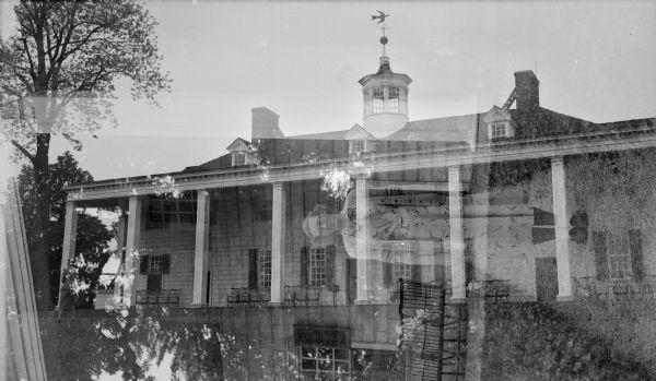Unintended multiple exposure of three images. Two are of the porch at George Washington's Mount Vernon (one vertical and distant, one horizontal and close-up). The other is a woman standing along a path in a wooded area. The cupola on the roof of Mount Vernon has a weather vane depicting a dove with an olive branch.