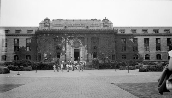 View from sidewalk towards the front of an ivy-covered Bancroft Hall at the Annapolis Navel Academy. Two cannons flank either side of the entrance, and a group of people are walking towards the front steps.