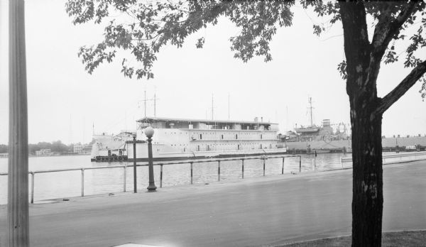 View across street towards several naval ships in the harbor at the United States Naval Academy. A sign near a lamppost reads: "Santee Basin."