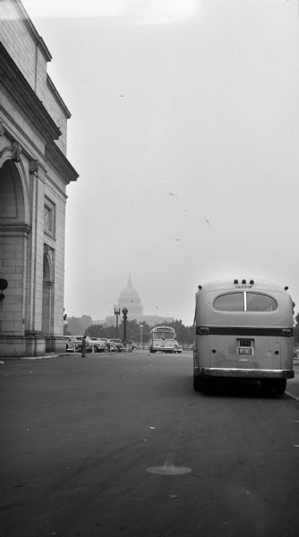 View from street towards the United States Capitol building. Several buses drive along the street with parked cars in a parking lot on the left.