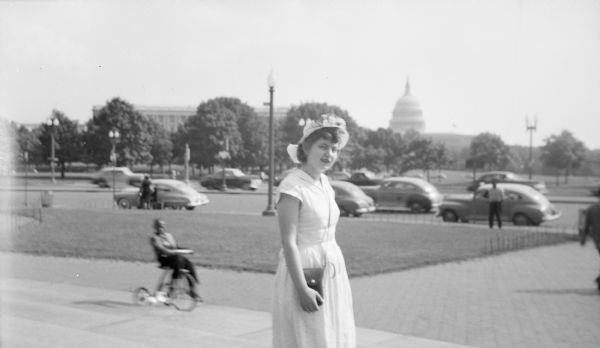 Shirley Krueger posing on stairs near a street, with the United States Capitol Building in the distant background. A child is riding a tricycle on the sidewalk on the left.