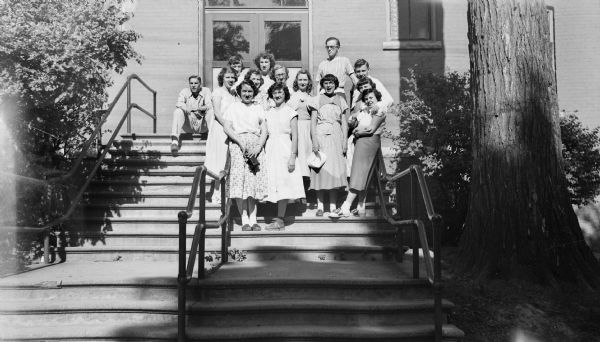 Group portrait of several women and men standing together on the stairs leading to the entrance of a building. One man sits separate from the group on the top step on the left.