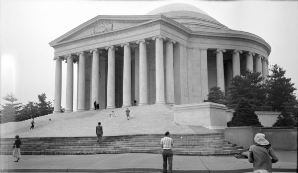 View of the Thomas Jefferson Memorial from the street, with people walking, sitting and standing on the sidewalk and on the stairs leading up to the memorial.