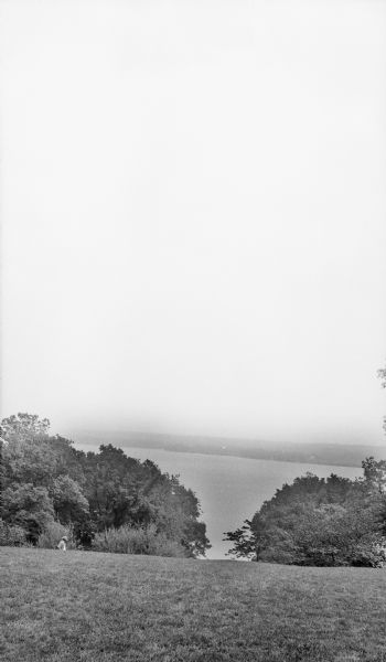View across lawn looking down from hill towards a body of water, with a far shoreline in the distance. Treetops are further down the hill. There is a man walking along the treeline below on the left.