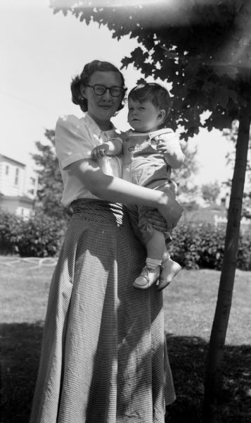 Portrait of a woman standing in a yard next to a maple tree, holding a child in her arms,