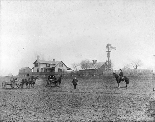 View of the Krueger family standing in a cleared field, with a view of their farm in the background. August Krueger is sitting with a dog in a wagon pulled by a team of two horses on the left, while Sarah and Mary Krueger are sitting together in a horse-drawn carriage. William Krueger is standing in the middle of the group holding a cane. Alexander Krueger is on the far right sitting on a horse.