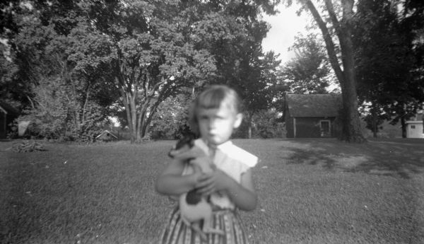 A young girl stands in the foreground holding a puppy in her arms. Behind her bordering the yard are trees and buildings.