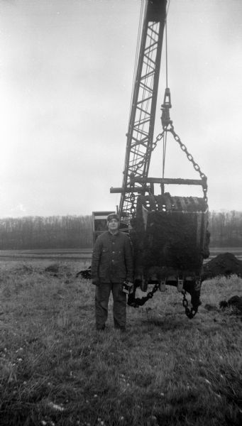 Robert Krueger posing in a field next to a drag bucket filled with dirt attached to a crawler crane. Edgar Krueger sits behind him in the cab of the crane. In the background is a forested hill.