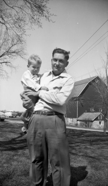 Robert Krueger standing in the yard holding his nephew, Glenn Oestreich. A barn and automobiles are in the background.