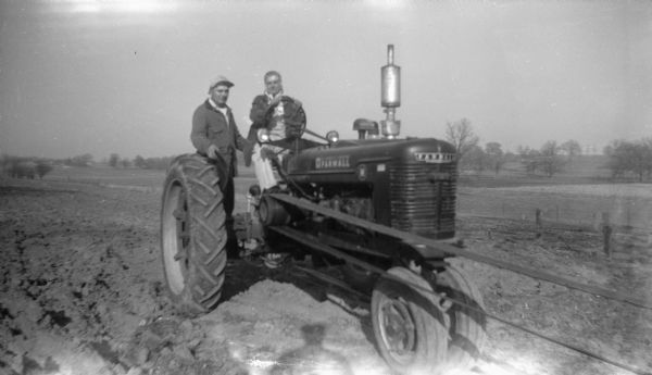 A couple posing on a Farmall Tractor in the middle of a plowed field. The woman sits in the driver's seat, while the man stands on the axle next to her. The tractor is set up to belt-drive equipment which is off-camera on the far right.