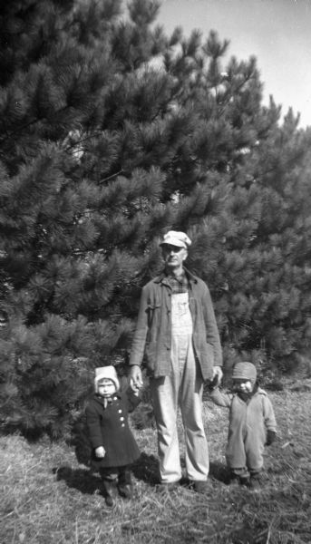 Edgar Krueger standing in front of a line of pine trees, with a young boy and girl on either side of him holding his hands.