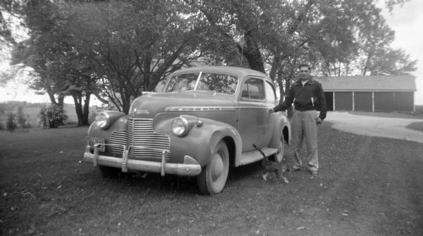 Robert Krueger stands near a driveway next to a Chevy automobile with his hand on the driver's side door knob. A cat is walking on the ground in front of him. In the background is a house and garage, and on the far left, trees and fields.
