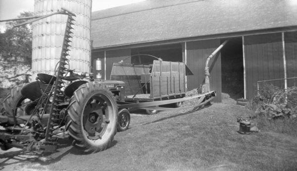 A large wagon and Farmall H tractor with an attached cultivator sitting next to a barn. The tractor is being used to belt-drive a blower that is running into the open barn door. A silo is next to the barn on the right.