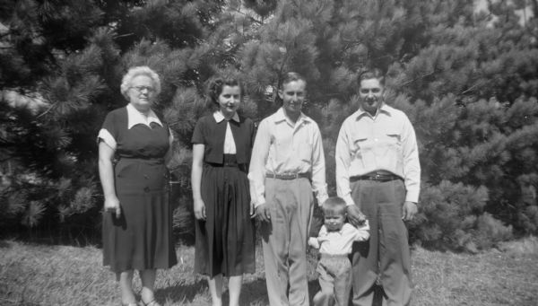 Group portrait of the Edgar Krueger Family standing in front of a line of trees. From left to right: Elna Krueger, Shirley Krueger Oestreich, Adelbert Oestreich, Glenn Oestreich, and Robert Krueger.