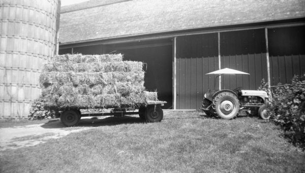 A wagon attached to a tractor is loaded with hay bales, stacked five high, and parked in front of an open barn door. A silo is next to the barn on the left.