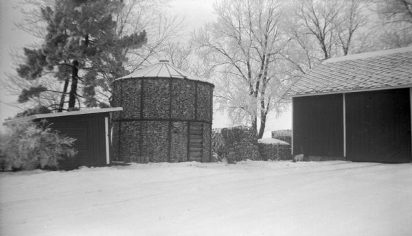 View across barnyard towards a full corn crib situated between a barn and small shed in front of a row of trees. A fresh blanket of snow covers the ground.