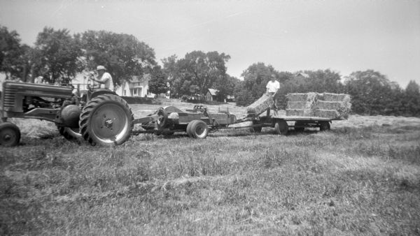 Left side view of man driving a John Deere tractor pulling a square baler attached to a wagon. Robert Krueger stands on the wagon behind the baler, stacking the hay bales coming off the baler. A farmhouse is on a low rise in the background.