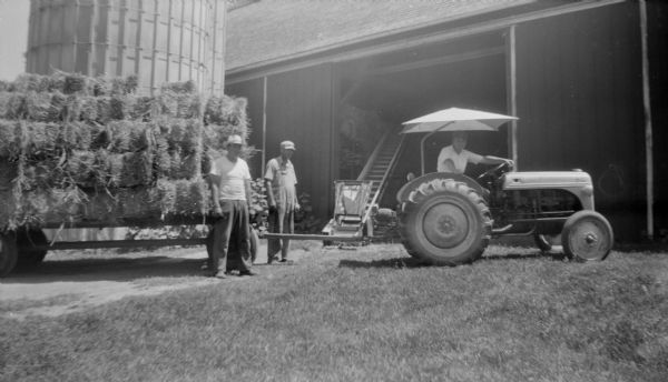 View across barnyard towards three men posing with freshly baled hay bales stacked on a wagon pulled by a small tractor. Robert Krueger sits under an umbrella on the tractor while his father, Edgar Krueger, stands in overalls near a conveyor. Another man stands in front of the wagon. The tractor and wagon are parked in front of an open barn door with a conveyor placed inside. A silo is next to the barn on the left.