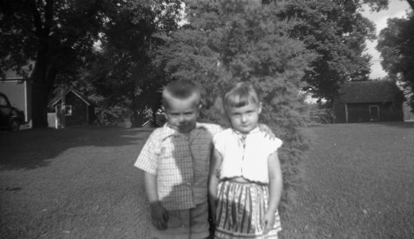 Outdoor portrait, with the shadow of Edgar over them, of Glenn Oestreich with his arm around the shoulder of a young girl. The two stand in front of a small tree in the Krueger's yard.